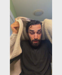 An adult man with dark hair and a beard smiling and holding a bottle of TRUBLOOM Hair Thicken Spray for instant volume.